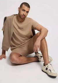 Beżowa koszula z krótkim rękawem z ciemnym nadrukiem, dopasowane szorty oraz kremowe sneakersy z czarnymi akcentami. Model pozując siedzi z szarym kurtką.