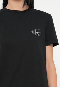 Μαύρο βαμβακερό T-shirt με στρογγυλή λαιμόκοψη και κοντά μανίκια. Εντυπωσιακό ασημί κείμενο γραφικών «Calvin Klein» με στυλιζαρισμένο λογότυπο «CK» στο στήθος.