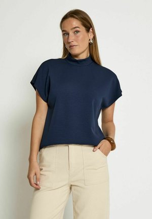 Navy blue short-sleeve top with a high neckline and a relaxed fit. Soft, smooth fabric with a minimalist design. Worn with beige high-waisted pants.