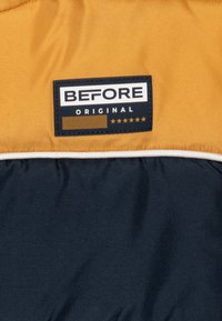 Veste bicolore composée de tissus jaune et bleu marine, avec un patch logo texturé indiquant "BEFORE ORIGINAL" et cinq étoiles. Couture blanche en accents.
