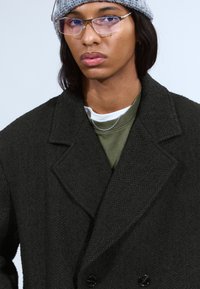 Donker visgraatjas met een dubbelzijdig ontwerp, gemaakt van textuurstof. Model draagt een grijze beanie over schouderlang haar.