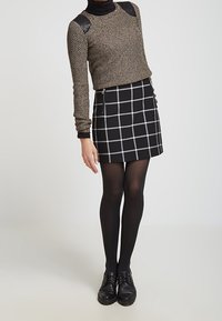 Falda a cuadros en blanco y negro combinada con un suéter de punto dorado con detalles en cuero negro en los hombros. La modelo lleva medias negras y zapatos.