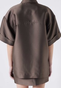 Brązowa satynowa oversize'owa koszula z krótkimi rękawami, o luźnym kroju i subtelnym połysku. Szwy na plecach i rozcięcia po bokach.