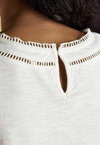 Valge puuvillane topp, millel on kitsas avaus tagaküljel, tekstureeritud serv ning peened lõike detailid kaeluses.