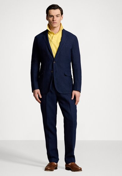 Laivastonsininen blazer, jossa on kaksi nappia ja kaksi taskua, päällä keltainen polo-paita. Housut sopivat blazeriin, ja ne on täydentänyt ruskeat kengät.