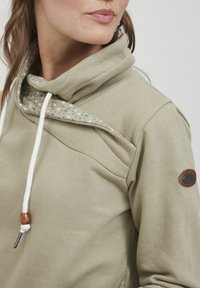 Felpa verde oliva con collo a cowl, con un interno decorato, lacci bianchi con dettagli in legno e una patch con logo sulla manica.
