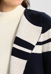 Stickad cardigan i en färgpalett av kräm och marinblått, med framträdande horisontella ränder och ett mjukt, strukturerat tyg.