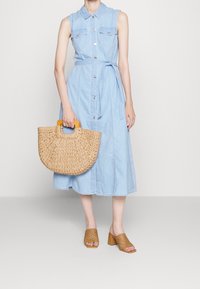 Jasnoniebieska sukienka dżinsowa z krótkimi rękawami, z guzikiem z przodu i paskiem do związania. Wykonana z plecionego słomianego materiału torebka w kształcie półksiężyca z drewnianym uchwytem. Brązowe, sandały na obcasie z otwartym noskiem.