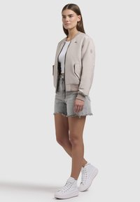 Lichtbeige bomberjack met zwarte rits en geribde manchetten, gedragen over een witte crop top. Gecombineerd met grijze denim shorts en witte high-top sneakers.