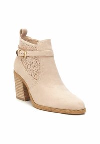 Botín de ante beige con puntera afilada, diseño lateral perforado y una correa decorativa con hebilla dorada. Tacón cuadrado con acabado texturizado.