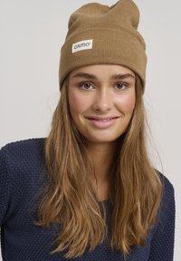 Gorro de punto color marrón claro con una etiqueta que dice "oxmo", que presenta un ajuste ceñido y un patrón texturizado. Modelo lleva un suéter texturizado azul marino.