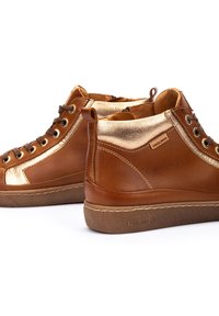 Bruna läder högstövlar med gyllene metalliska detaljer, rund tå, snörning på framsidan och gummisulor. Varumärket är synligt på sidan.