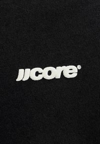 Λευκό κεντημένο κείμενο "JJ core" σε μαύρο υφασμάτινο φόντο με μικρό σύμβολο καταχωρημένου εμπορικού σήματος.