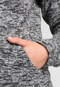 Mano parzialmente inserita nella tasca anteriore di un indumento a maniche lunghe in tessuto grigio e nero mélange.