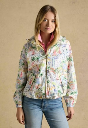 Junge Frau trägt eine bunte Zip-up-Jacke mit tropischem Segelboot- und Palmbaum-Motiv, kombiniert mit hellblauen Jeans.