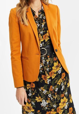Vrouw draagt een oranje blazer over een zwart bloemenjurkje met gele, witte en oranje bloemen, in de taille aangesnoerd met een brede zwarte riem.