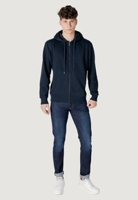 Felpa con zip di colore navy e cappuccio con cordino, abbinata a jeans blu e sneakers bianche. Il tessuto appare liscio e informale.