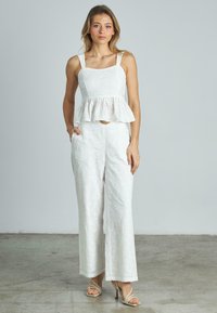 Weißes Peplum-Top mit quadratischer Ausschnitt und filigranem Blumendesign. Kombiniert mit weißen, weit geschnittenen Hosen, die auch seitliche Taschen haben.