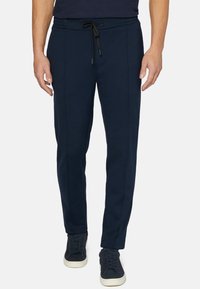 Man i mörkblå joggerbyxor med dragsko och matchande mörkblå sneakers, står med armarna avslappnat längs sidorna.