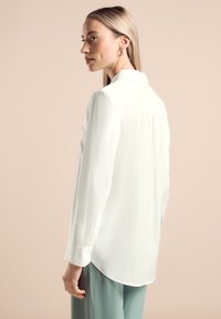 Camisa blanca de manga larga con cuello, de tejido suave, con detalles de pliegues en la parte posterior, combinada con pantalones verde claro. Textura suave, diseño casual.