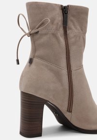 Bottines en suède beige avec un talon en bois brun. Dotées d'une fermeture éclair sur le côté et d'un accent noué en haut. Texture lisse et design ajusté.