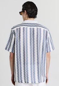 Mann in einem kurzärmeligen Hemd mit vertikalen Zickzackstreifen in Weiß, Blau und Grau, von hinten vor einem einfarbigen Hintergrund betrachtet.