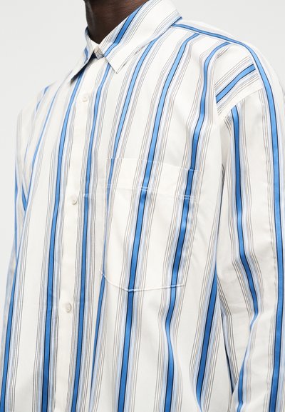 Άνδρας που φοράει λευκό πουκάμισο με κουμπιά, με κάθετες ρίγες σε μπλε, μαύρο και γκρι χρώμα και με τσέπη στο στήθος στην αριστερή πλευρά.
