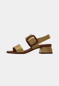 Χρυσό μεταλλικό σανδάλι με ανοιχτά δάχτυλα, με τακούνι block, φαρδιά λουράκι στον αστράγαλο και μεγάλη αγκράφα από χελώνα, σε απλό φόντο.