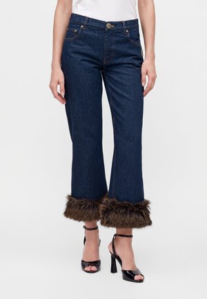 Jean court bleu foncé avec bordure en fourrure marron aux poignets, porté avec des sandales noires à talons hauts et un haut blanc.