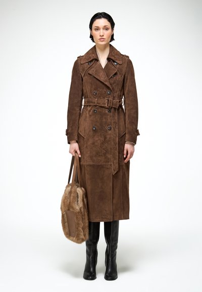 Vrouw staand in een bruine suède double-breasted trenchcoat, met een grote, harige bruine handtas, gedragen met zwarte kniehoge laarzen.