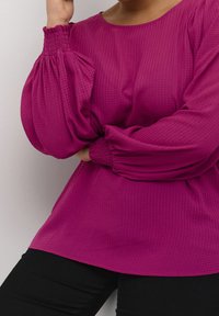 Femme portant un blouse magenta texturé avec des manches longues bouffantes et des poignets froncés, associée à un pantalon noir devant un mur uni.