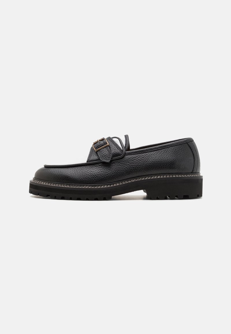 Mocassin en cuir noir avec une finition texturée, présentant une boucle décorative, une semelle basse avec des crampons de traction et des détails de surpiqûres contrastants.
