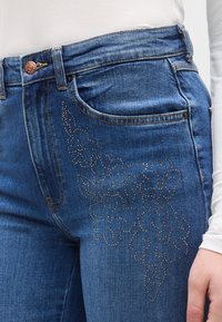 Μπλε τζιν με εφαρμοστή σχεδίαση, διακοσμημένο με κεντήματα λουλουδιών σε μεταλλική κλωστή στο αριστερό μηρό. Περιλαμβάνει μπροστινή τσέπη και χαλκοκόκκινο κουμπί.