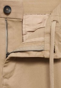 Pantalon beige avec une fermeture éclair partiellement ouverte, une fermeture bouton et un cordon beige tressé passé dans une boucle en tissu.