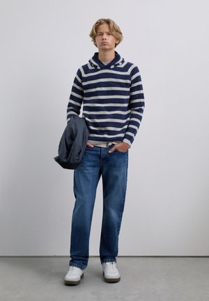 Jonge man in een marineblauw en grijs gestreepte hoodie, blauwe jeans en witte sneakers, met een donker jack in zijn linkerhand.