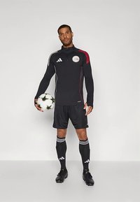 Haut de survêtement athlétique noir avec des accents rouges, short noir et chaussettes rayées. Tenant un ballon de football, avec un logo Adidas sur les vêtements.