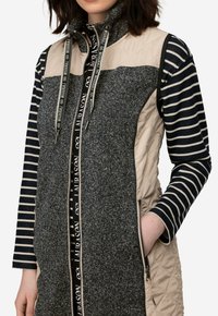 Kleurgeblokte vest met grijs gebreid materiaal en beige gewatteerde panelen. Heeft een hoge kraag, ritssluiting en gestreepte lange mouwen.