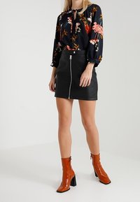 Zwarte faux leren rok met ritsdetail aan de voorkant, gecombineerd met een donkerbloemige blouse met lange mouwen en een striknek en wijd uitlopende mouwen. Oranje hakken maken de look compleet.