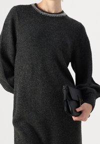 Pull en laine gris foncé à manches longues et coupe ample, avec un col orné de perles. Une pochette noire est tenue dans la main droite.