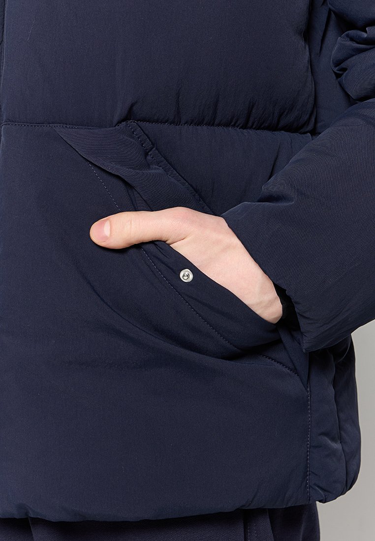 Veste matelassée bleu marine avec une poche latérale à fermeture à bouton-pression et texture matelassée, fabriquée à partir d'un tissu léger et résistant à l'eau.