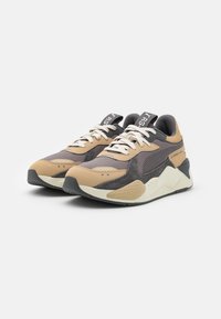 Zapatillas deportivas que presentan una combinación de materiales marrones, grises y beige con un diseño texturizado, cordones planos y una lengüeta acolchada.