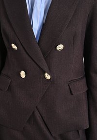 Blazer marrone a doppio petto in herringbone con bottoni dorati, rever a punta e due tasche frontali. Camicia a righe blu chiaro sotto.