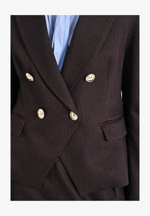 Doppelt gefütterte braune Fischgrätblazer mit goldenen Knöpfen, Revers und zwei Fronttaschen. Darunter ein hellblau gestreiftes Hemd.