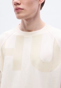 Ανοιχτό μπεζ φούτερ με μεγάλο, ημιδιαφανές λογότυπο «HU». Διαθέτει στρογγυλή λαιμόκοψη και μανίκια ράγκλαν σε απαλό, υφασμάτινο ύφασμα με υφή.