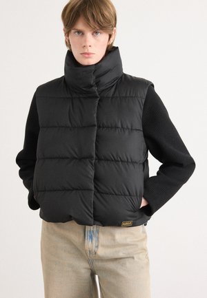 Zwarte puffer gilet met hoge kraag, geborduurd patroon en ritssluiting, gecombineerd met een zwart gestructureerd trui met lange mouwen en lichtgekleurde spijkerbroek.