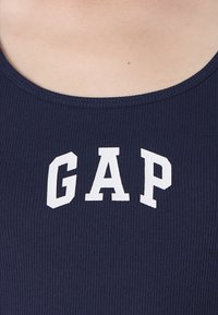 Majică din material ribbed de culoare bleumarin, cu inscripția albă "GAP" pe piept, prezentată purtată pe partea superioară a corpului unei persoane.