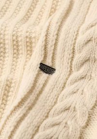 Kremfarvet kabelstrikstof med et lille sort mærke med teksten "SUPERDRY" syet ind i sømmen.