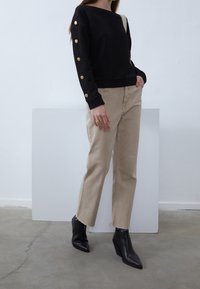 Maglietta nera a maniche lunghe con texture e bottoni dorati, abbinata a pantaloni dritti beige chiaro. Stivaletti neri con tacco e punta appuntita.
