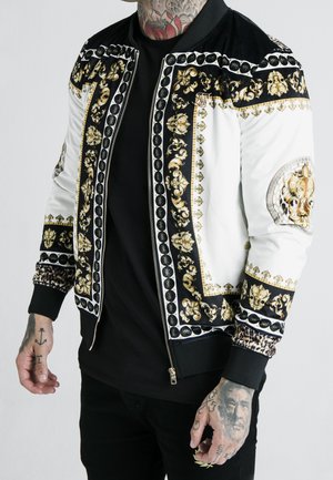 Mężczyzna w czarno-białej kurtce bomber z misternymi złotymi wzorami barokowymi i godłem lwa na rękawie, czarną koszulą i wytatuowanymi dłońmi.