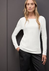 Top blanco de manga larga con escote cuadrado, combinado con pantalones gris oscuro. La tela tiene una textura suave y se ajusta en la cintura.